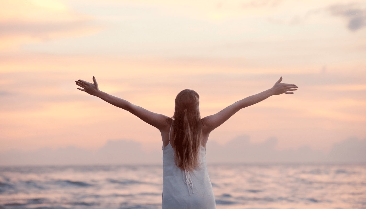 A woman in a white dress is standing on the beach with her arms outstretched.