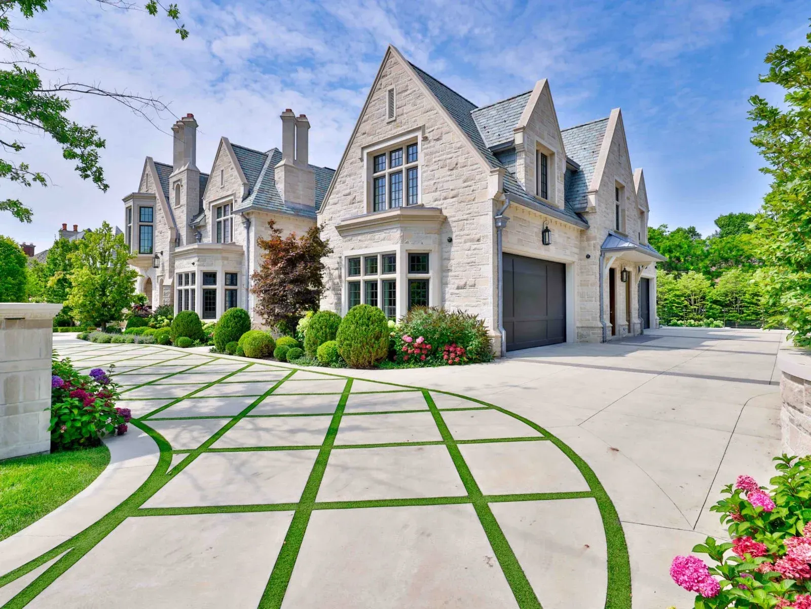 Luxury stone mansion with gray garage, curved driveway, and landscaped garden under a blue sky
