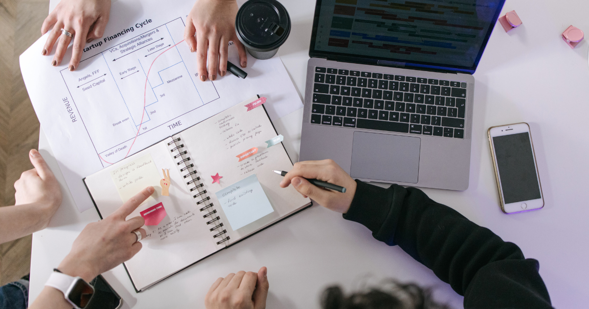 Team meeting at a desk with laptop, notebooks, charts, and hands pointing at notes.