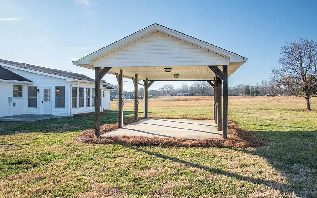 A white carport is sitting in the middle of a grassy field in front of a house.
