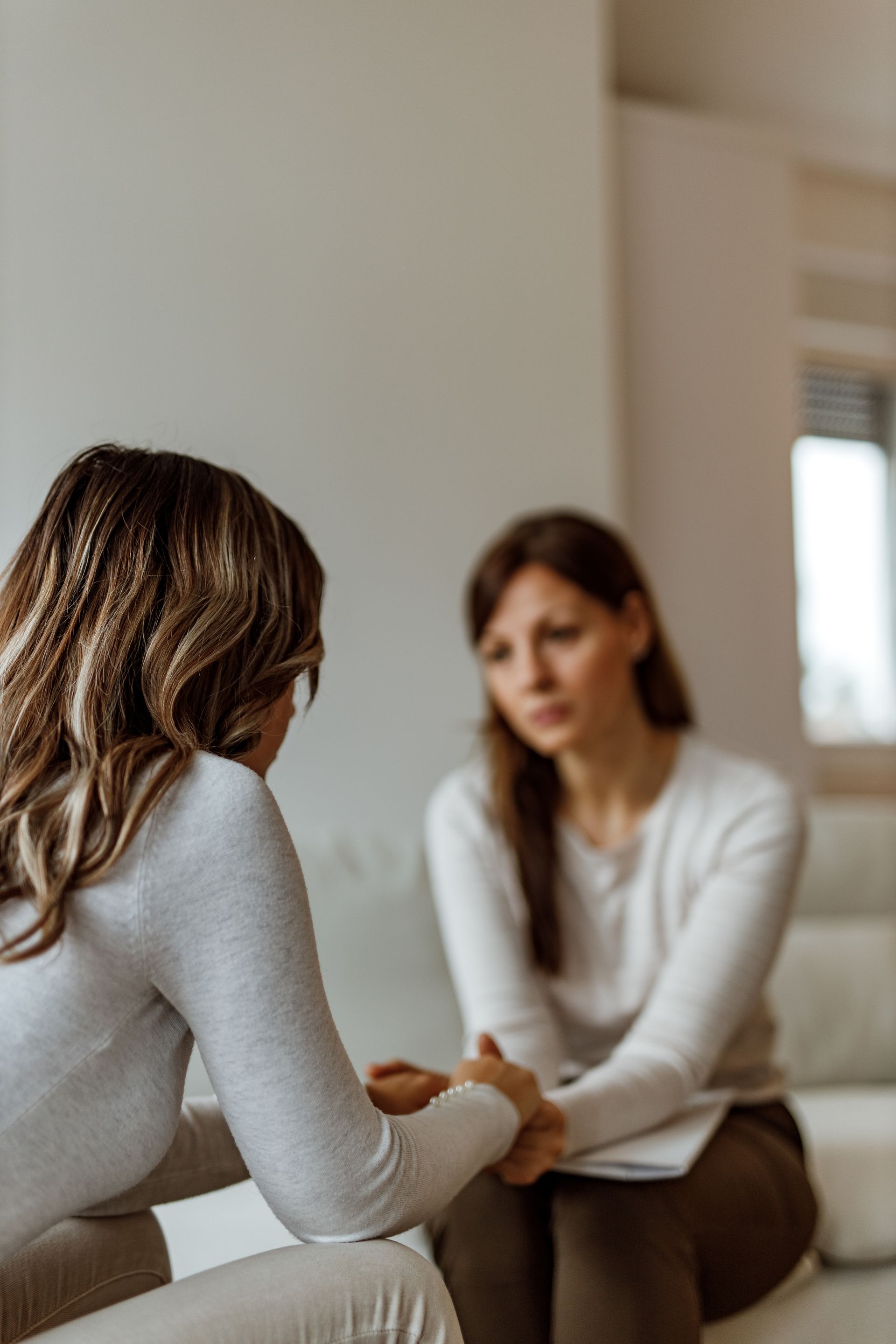 Woman comforting another woman in a light-filled room; one woman holds the other's hands, expressing concern. Woman comforting another woman in a light-filled room; one woman holds the other's hands, expressing concern.