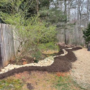 Curved garden bed with dark mulch, white stones, and young shrubs along a wooden fence beside a gravel path