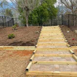 Wooden staircase walkway up a landscaped hillside with mulch, shrubs, and trees behind a fence