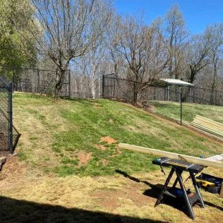 Sunny backyard with a grassy slope, trees, fencing, and sawhorses in the foreground