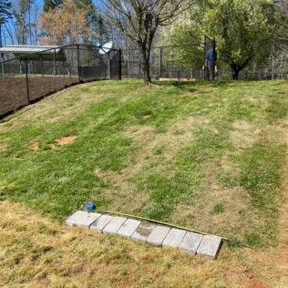 Grassy sloped yard with a chain-link fence and trees, plus a small row of pavers at the bottom.
