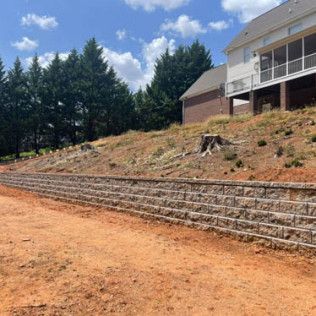 Stone retaining wall beside a dirt slope and house, with trees and blue sky in the background
