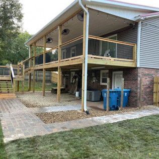 Raised deck attached to a house, with wooden railing, stairs, and blue bins underneath beside a brick foundation.