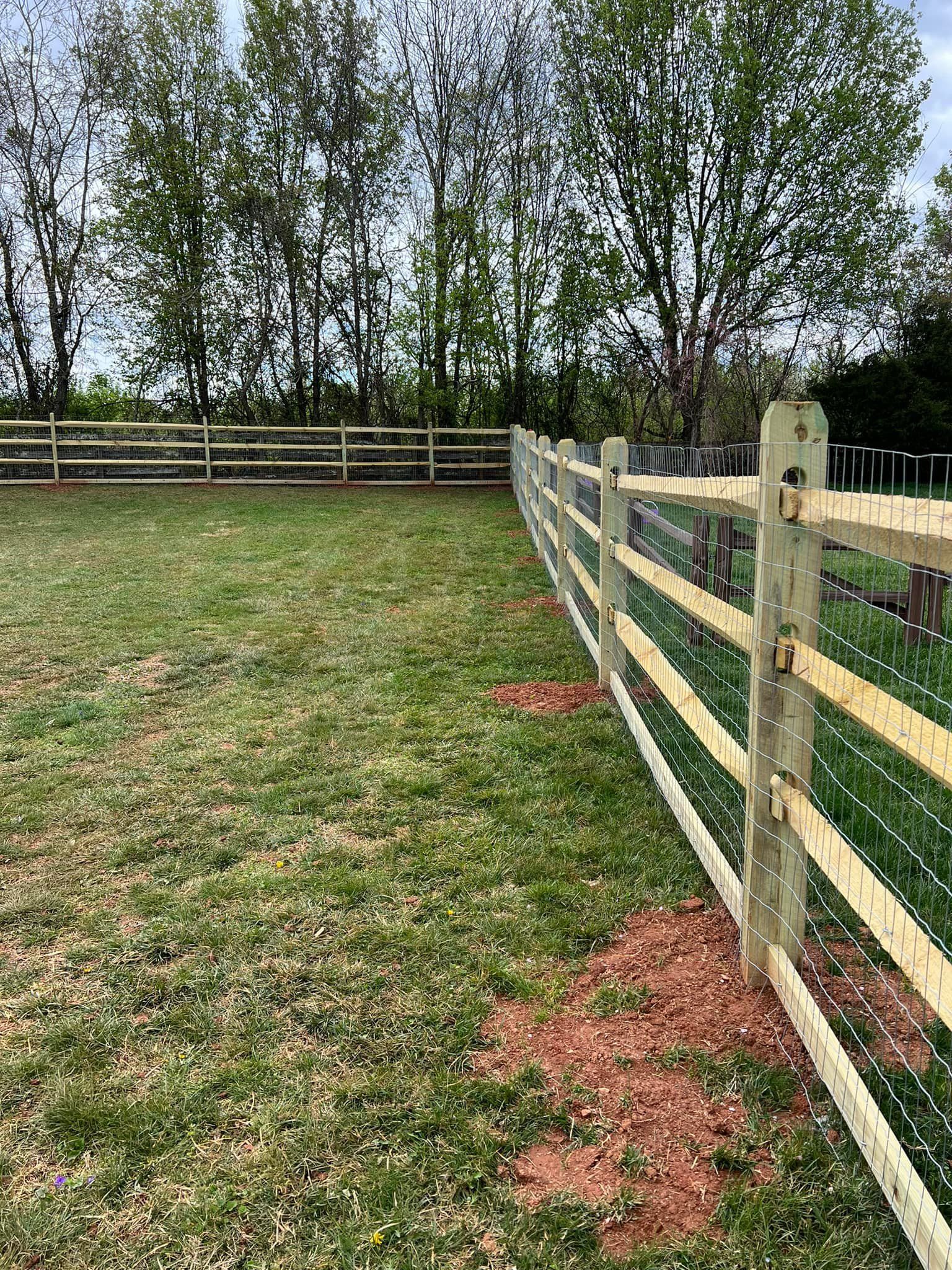 Wooden fence along a grassy yard with trees in the background and red dirt at the base.