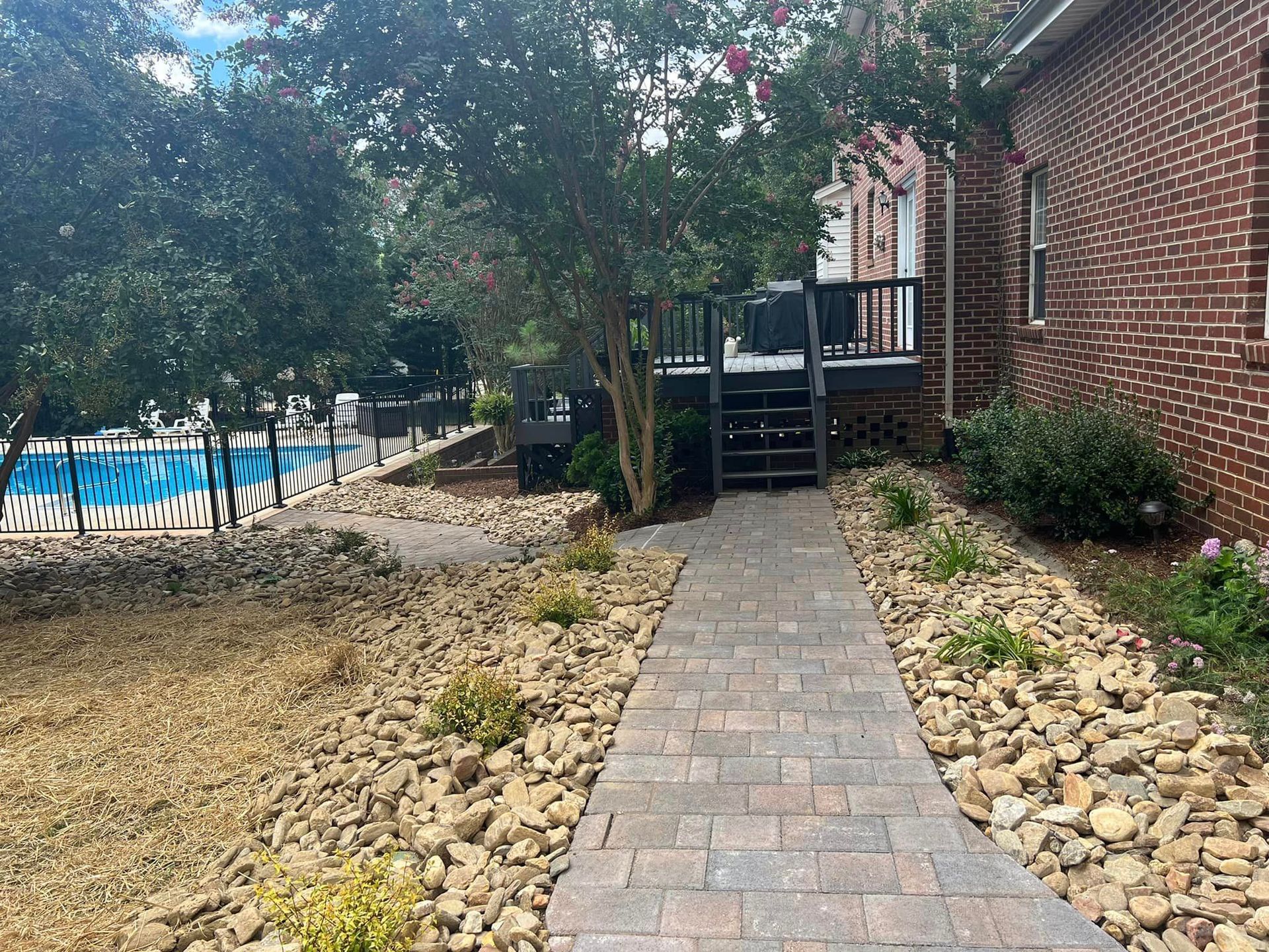 Brick paver path beside a house leading to a deck, with rocks, trees, and a backyard pool nearby.