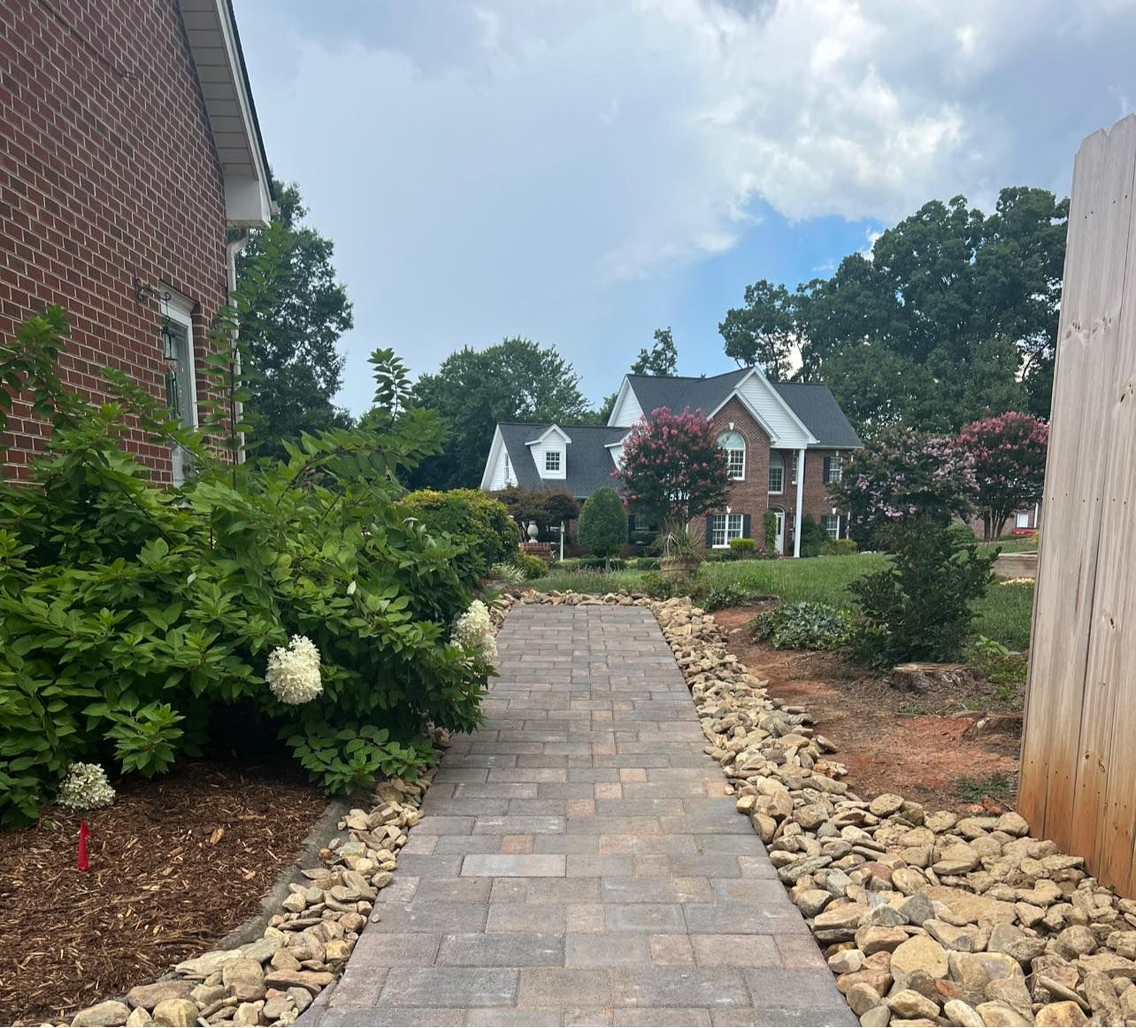 Stone paver walkway between houses, bordered by rocks and shrubs, leading to a landscaped front yard.