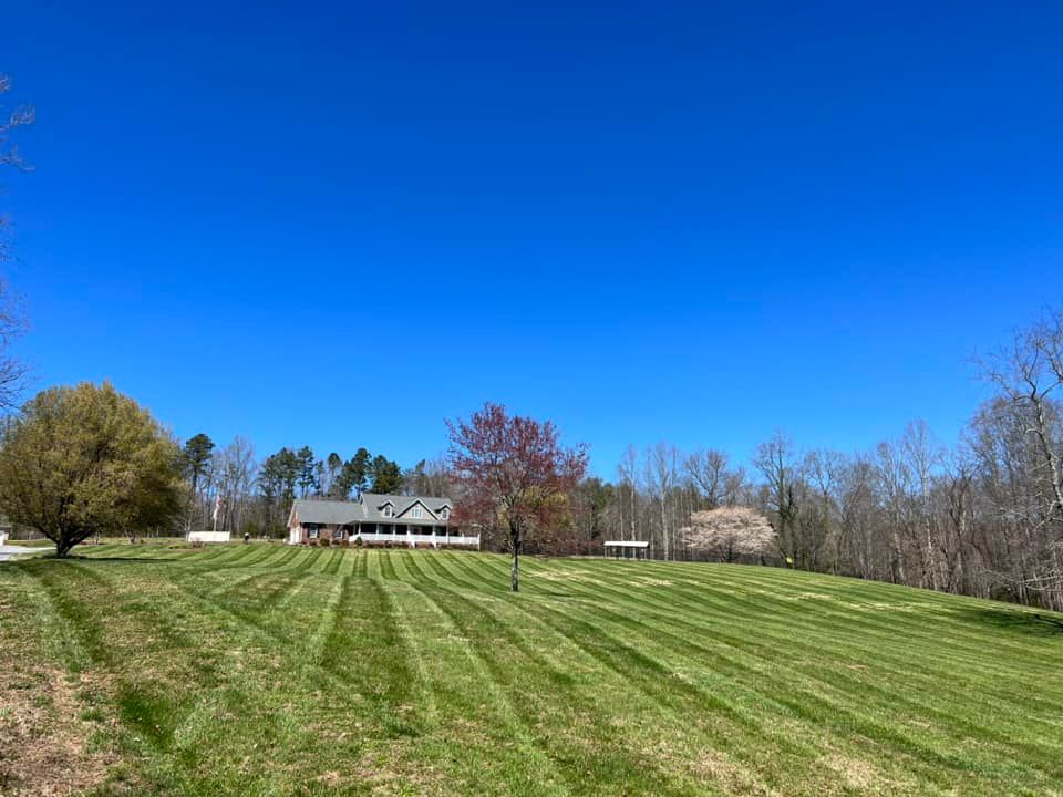 Wide grassy field with mowed stripes, trees, and a house under a clear blue sky.