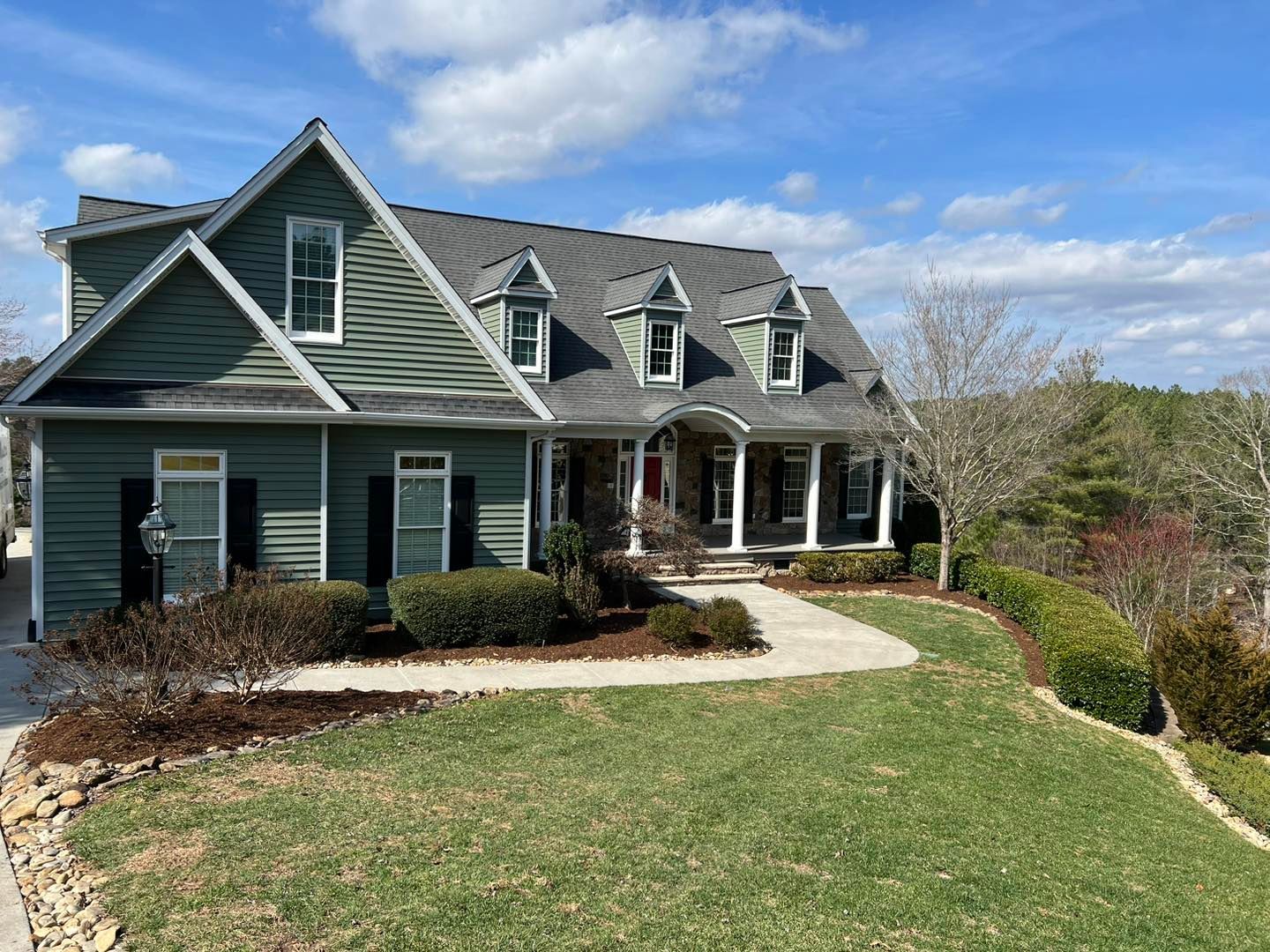 Green house with gabled roof, white trim, front porch, and curved walkway on a grassy lawn under blue sky