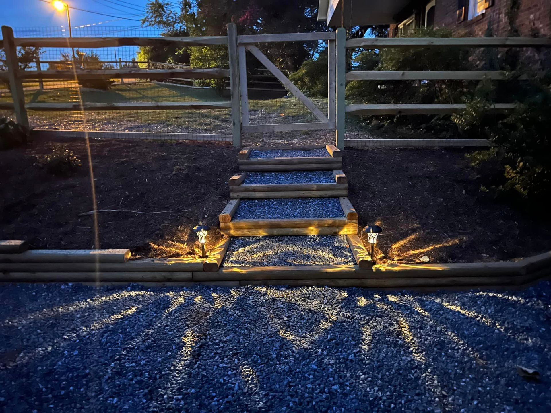 Lit steps leading to a metal farm gate at dusk, with gravel foreground and a wooden fence behind.