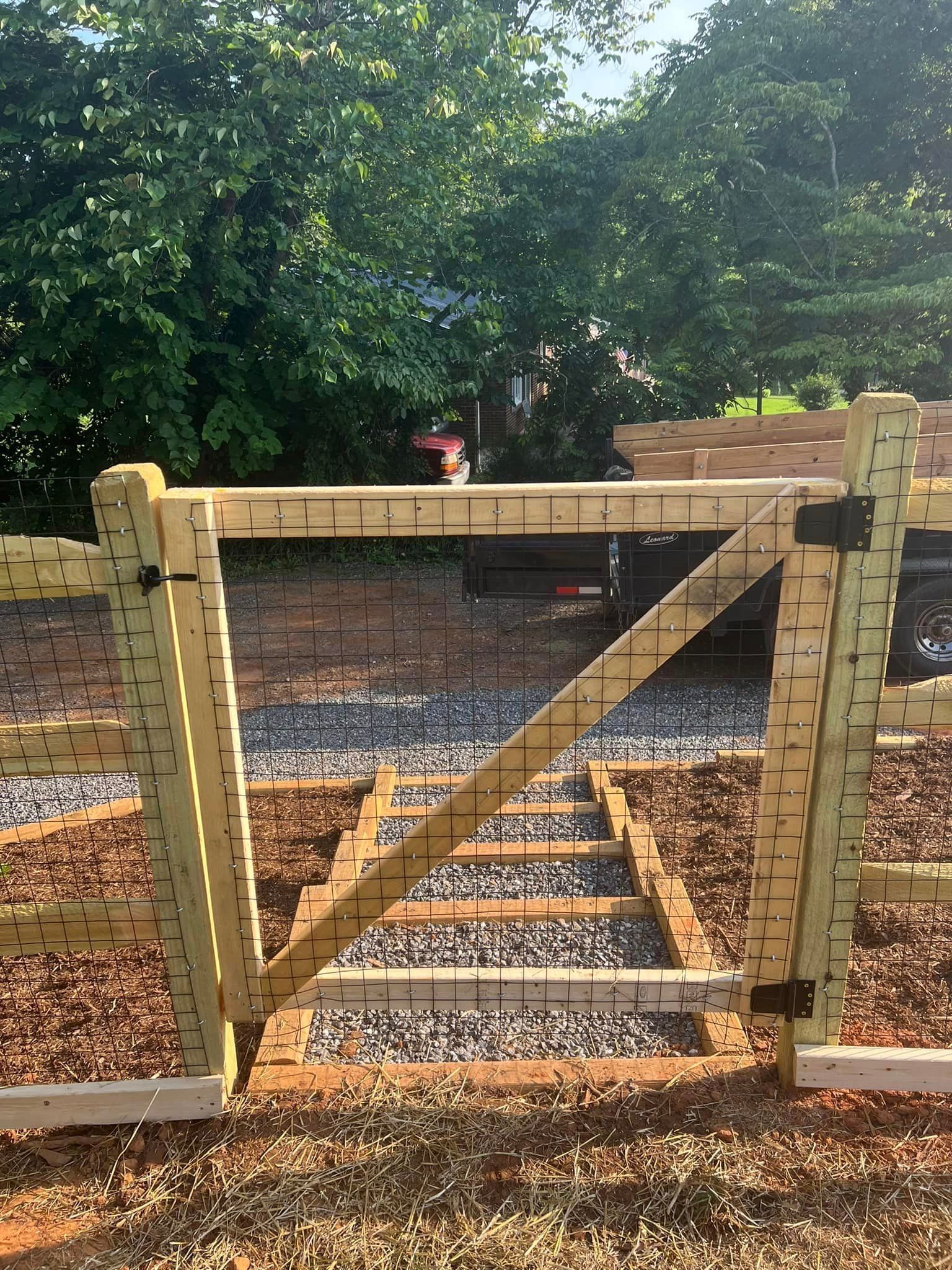 Wooden farm gate with diagonal brace, closed across a gravel path under leafy trees.