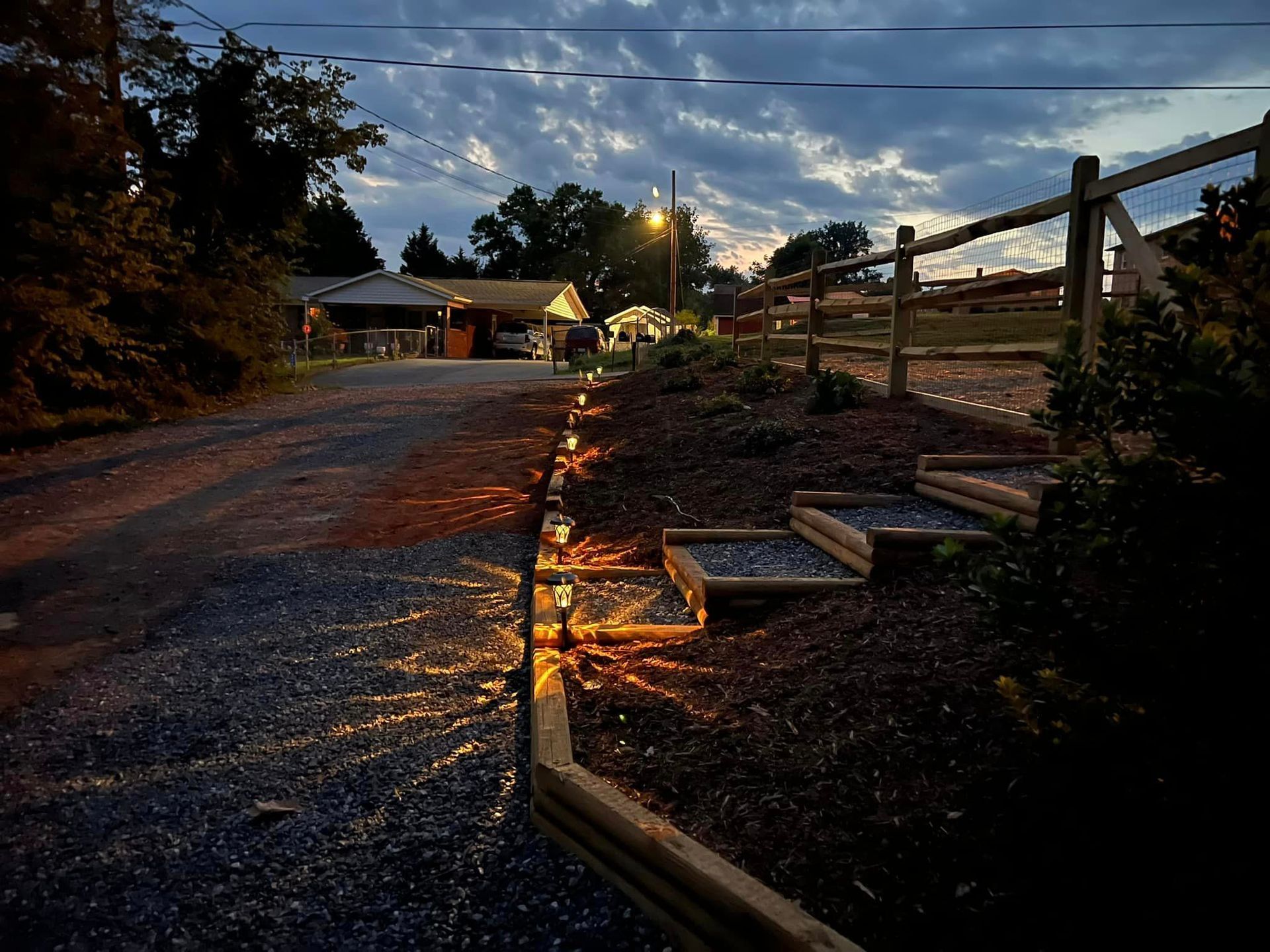 Dusk over a rural gravel path with a wooden fence, streetlights, and houses in the distance