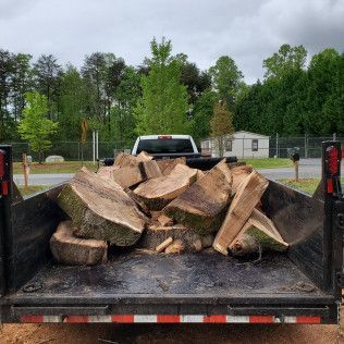 Flatbed trailer loaded with cut firewood logs in a parking lot, with a pickup truck and trees behind it