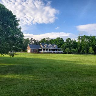 Large house with a porch in a green field under a blue sky, framed by trees