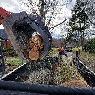 Excavator claw lifting a cut tree trunk from a trailer, with a person standing nearby on a driveway.