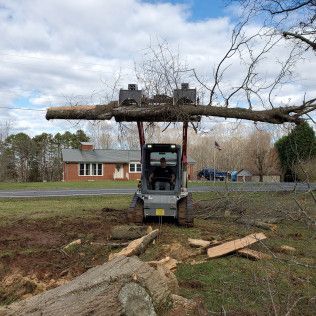 Small excavator lifting a large tree trunk in a muddy yard near a house