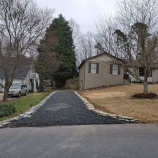 Residential driveway with fresh black gravel leading to a house in a wooded suburban yard on a cloudy day