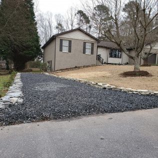 Gravel driveway leading to a tan house with bare trees and a white garage on a cloudy day