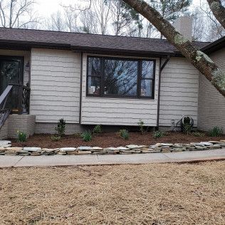 Single-story beige house with a large front window, stone border, and leafless tree in the yard