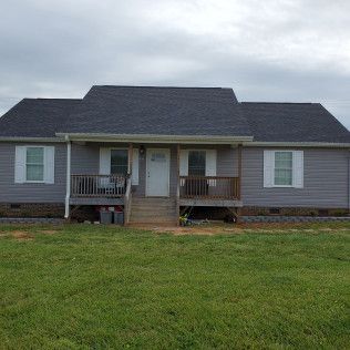 Gray house with a small front porch and steps, set behind a wide green lawn under cloudy skies