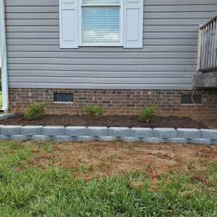House exterior with gray siding, brick foundation, small shrubs, and a stone-bordered mulch bed along the wall