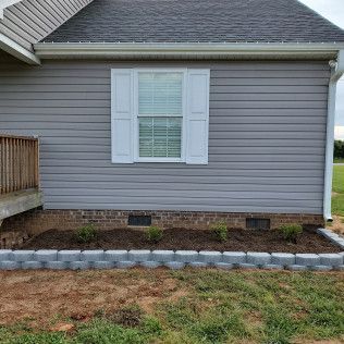 Gray house siding with a white shuttered window, bordered by a stone-edged flower bed and patchy grass.