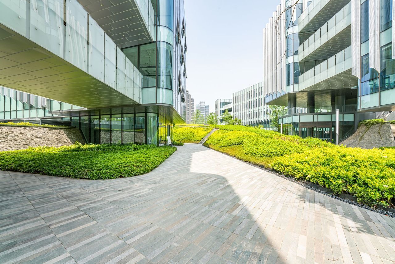 Modern office courtyard with curved glass buildings, gray paved walkway, and bright green landscaping