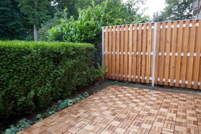 Small patio with patterned wooden pavers, a tall green hedge, and a slatted wooden fence.