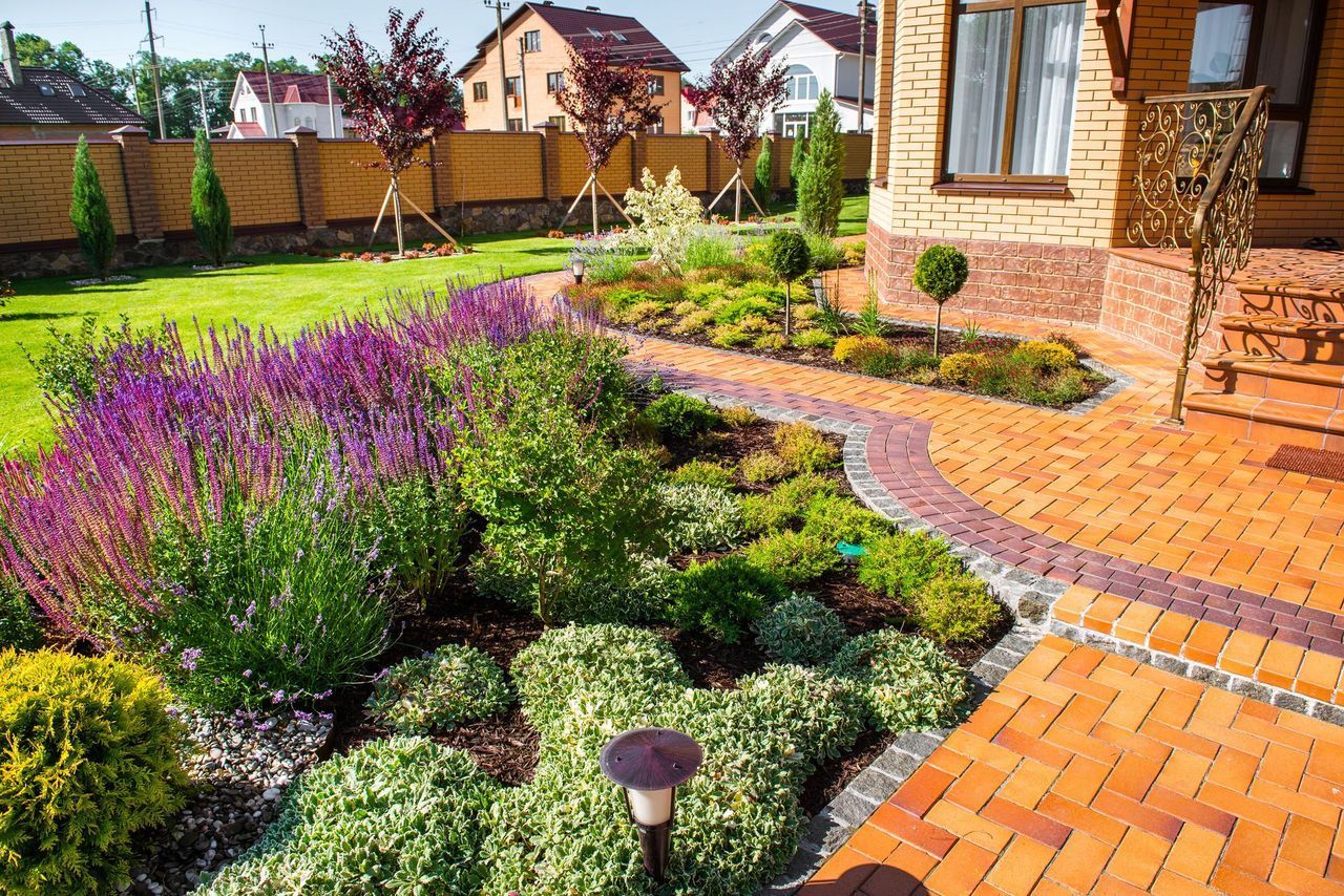 Colorful landscaped garden with purple flowers, shrubs, and a brick walkway beside a house