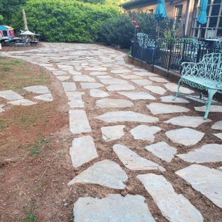 Stone pathway through a garden beside a house, with a metal bench and outdoor seating.