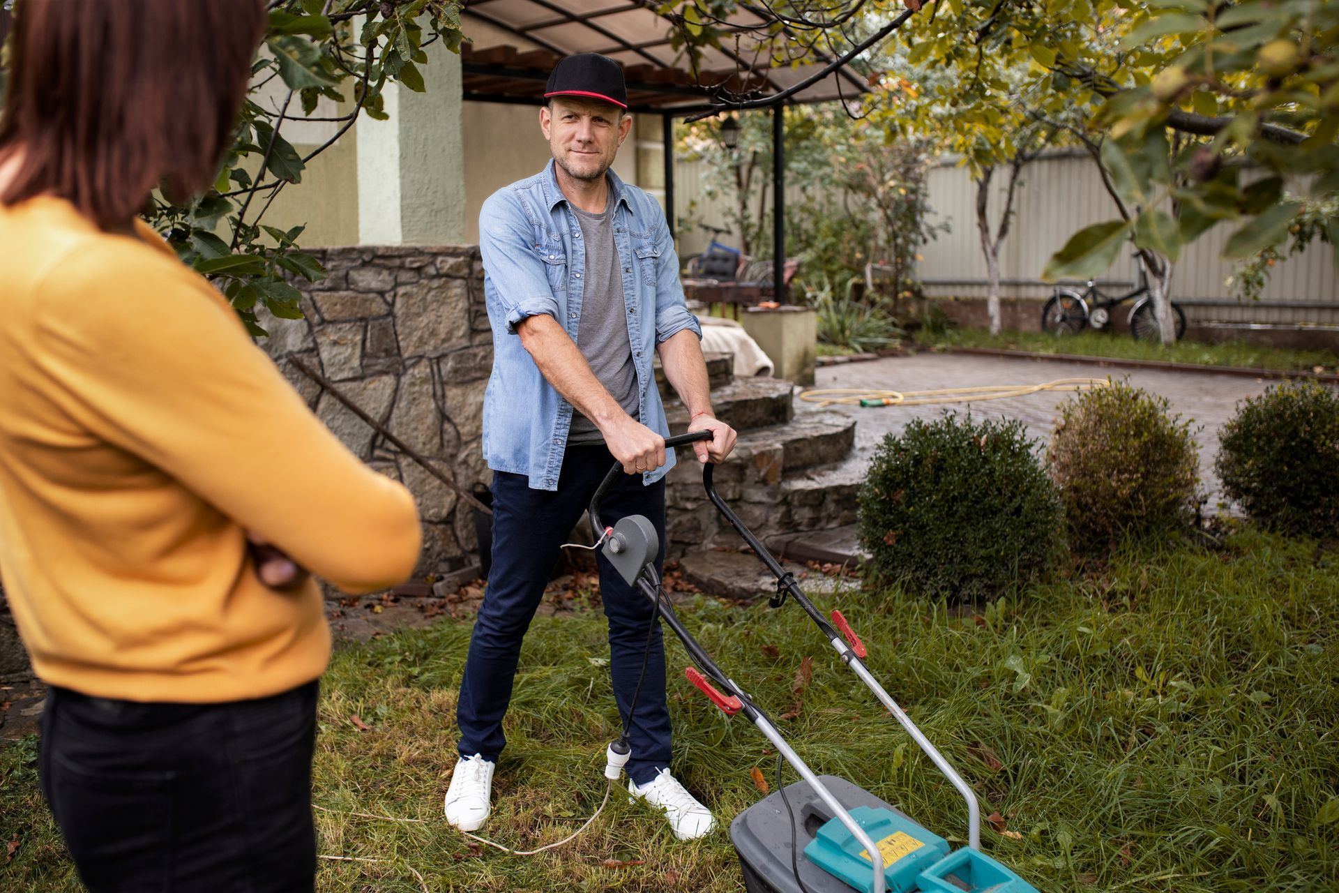 Man mowing a lawn, woman watches with arms crossed, in a backyard.
