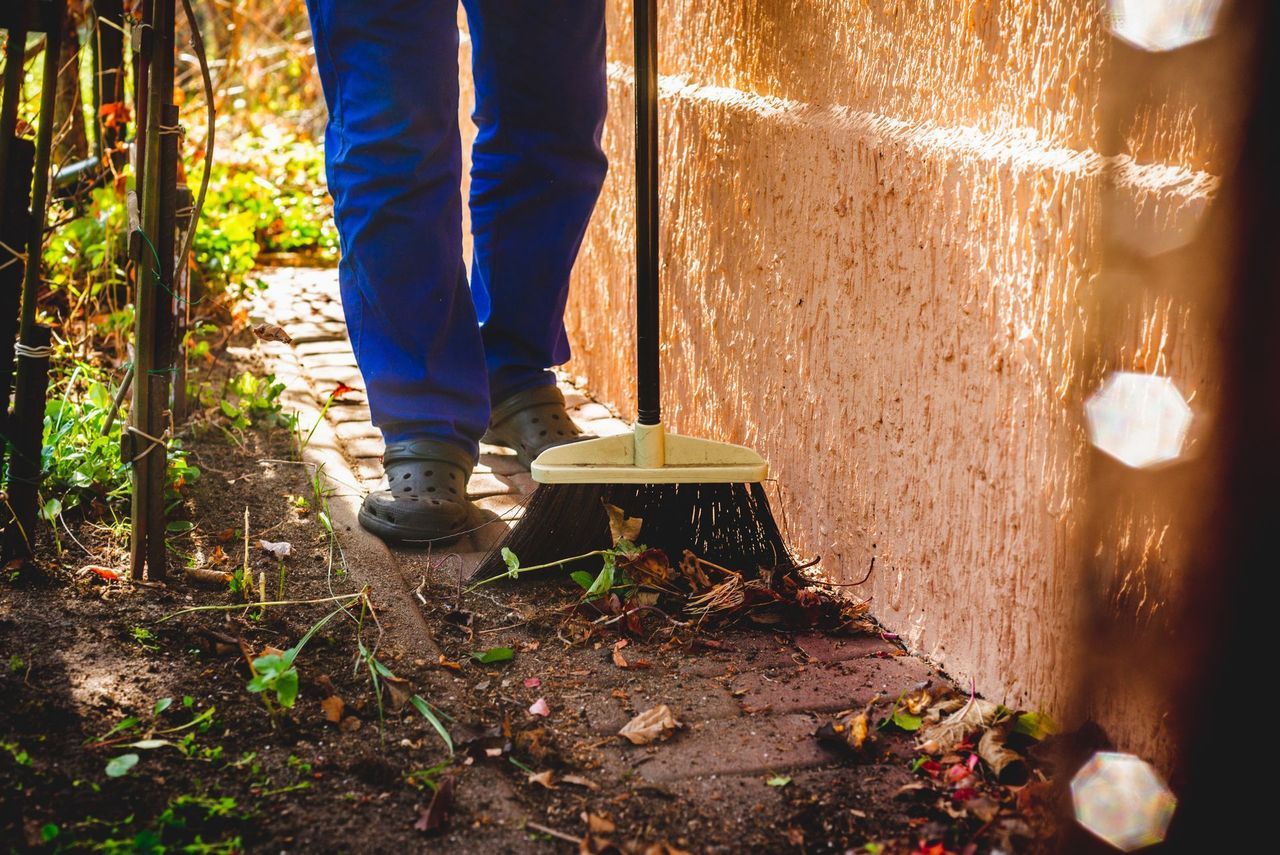 Person sweeping dirt beside a wall on a sunlit outdoor path