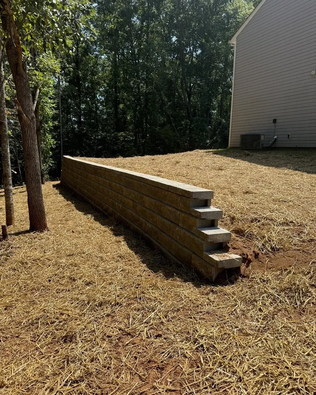 Stacked wooden retaining wall on a sloped, mulched yard beside a house and trees.
