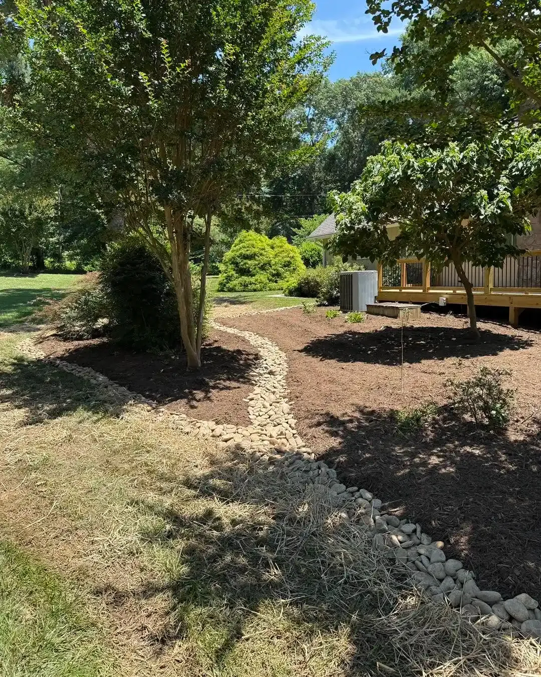 Shaded backyard with trees, mulch beds, and a curved stone border along a dirt path