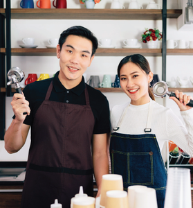 Two smiling baristas in aprons holding kitchen tools behind a café counter