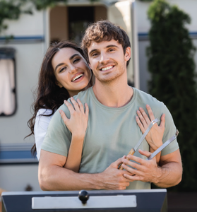 Smiling couple embracing outdoors behind a grill, with the woman leaning on the man’s shoulder