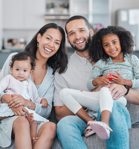 Smiling family of four sitting on a couch in a bright living room, holding two young children
