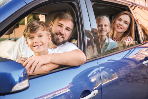 Family smiling and leaning out of a blue car window, with a child visible in the back seat.