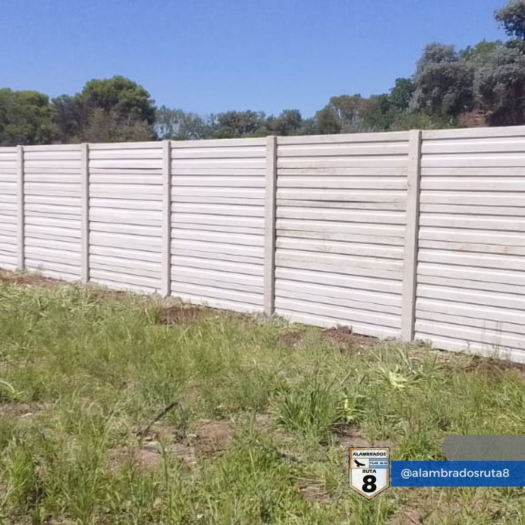 Valla de listones horizontales de hormigón en un campo de hierba bajo un cielo azul.