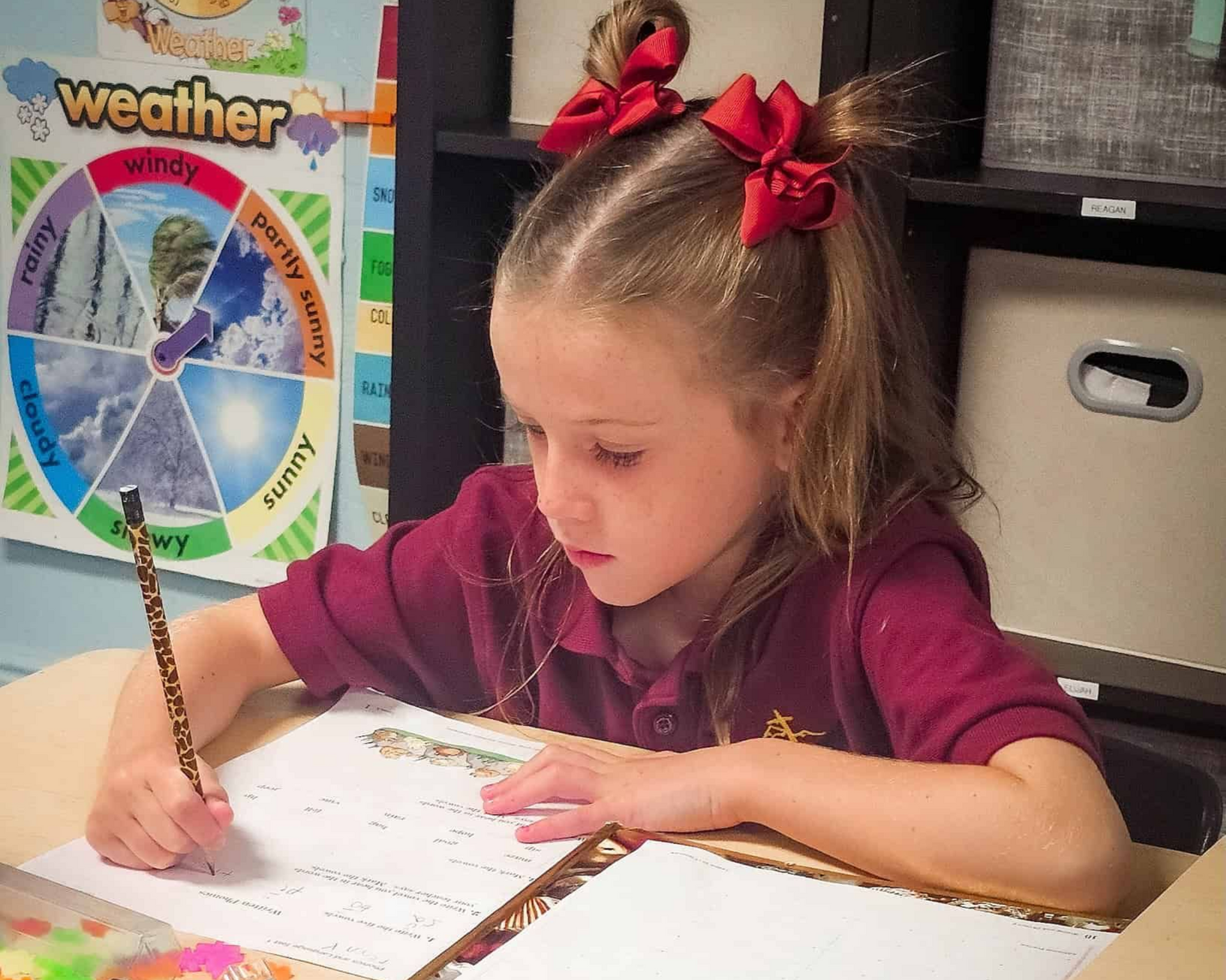 Child writing at a desk with a pencil, near a colorful weather chart