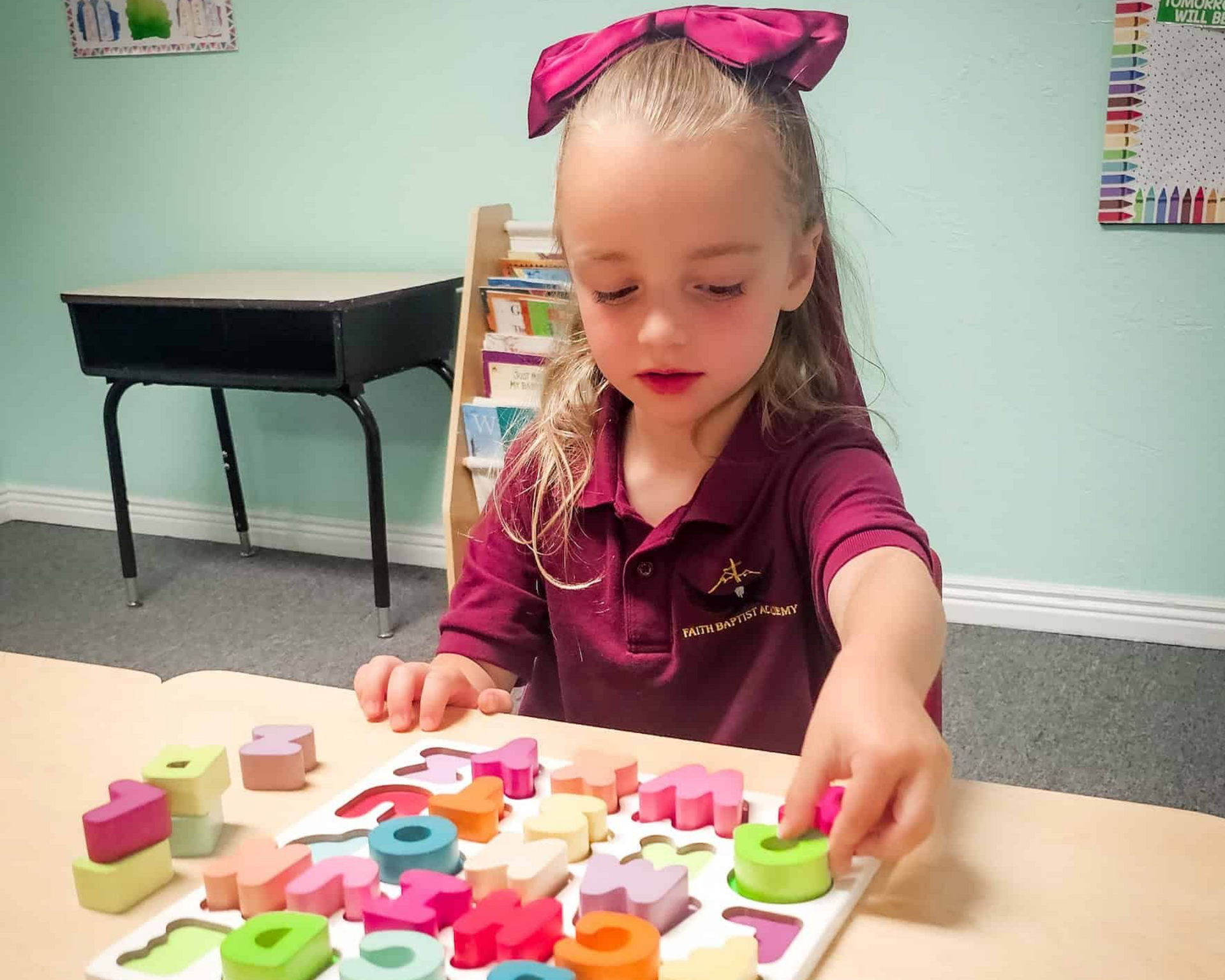 Child in maroon shirt reaching for colorful shape puzzle pieces at a table in a classroom