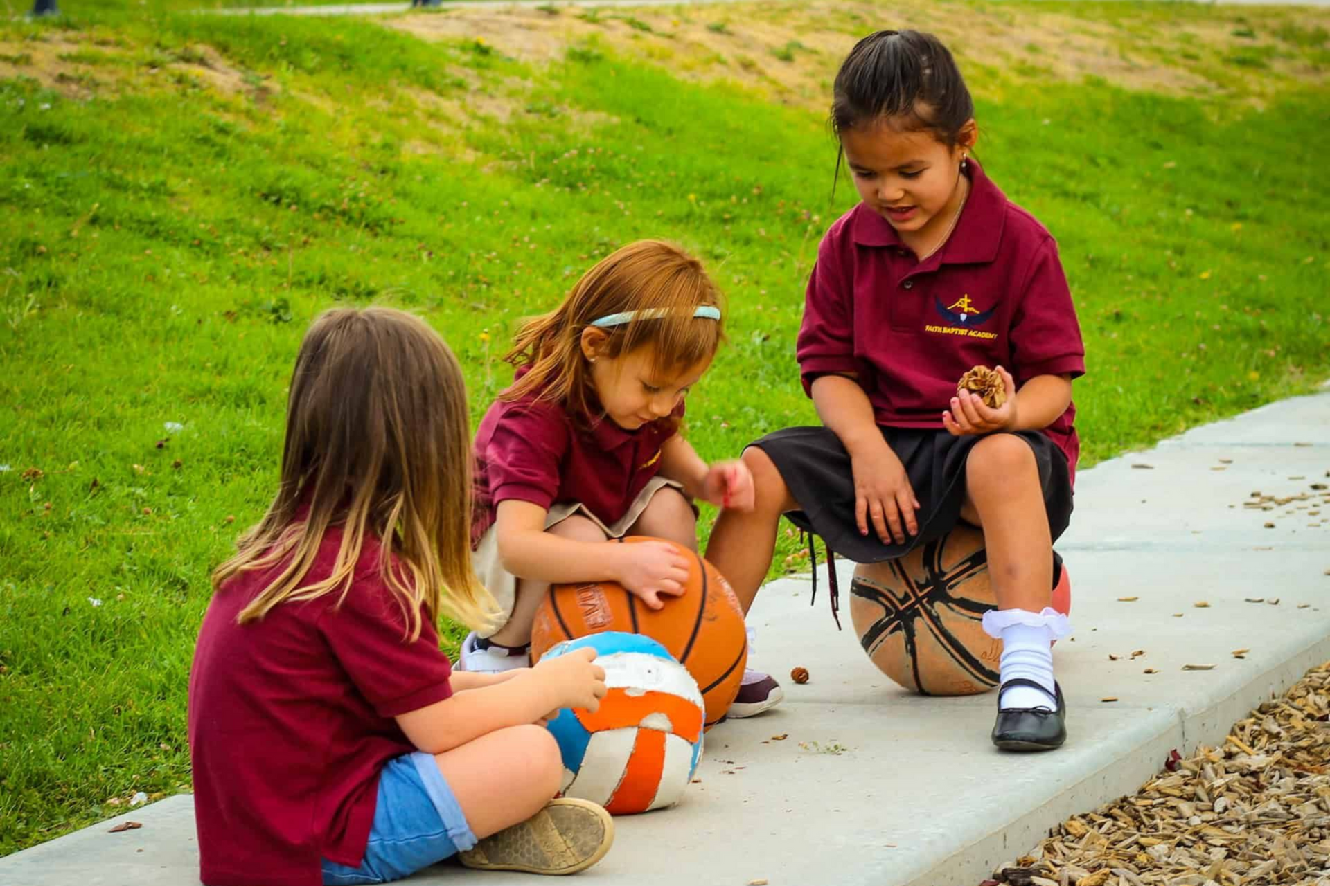 Three children in maroon shirts sit on a curb playing with an orange-and-white ball on a grassy sidewalk.