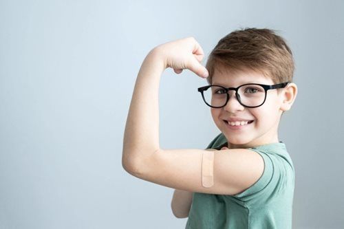 A young boy with glasses and a bandage on his arm is flexing his muscles.