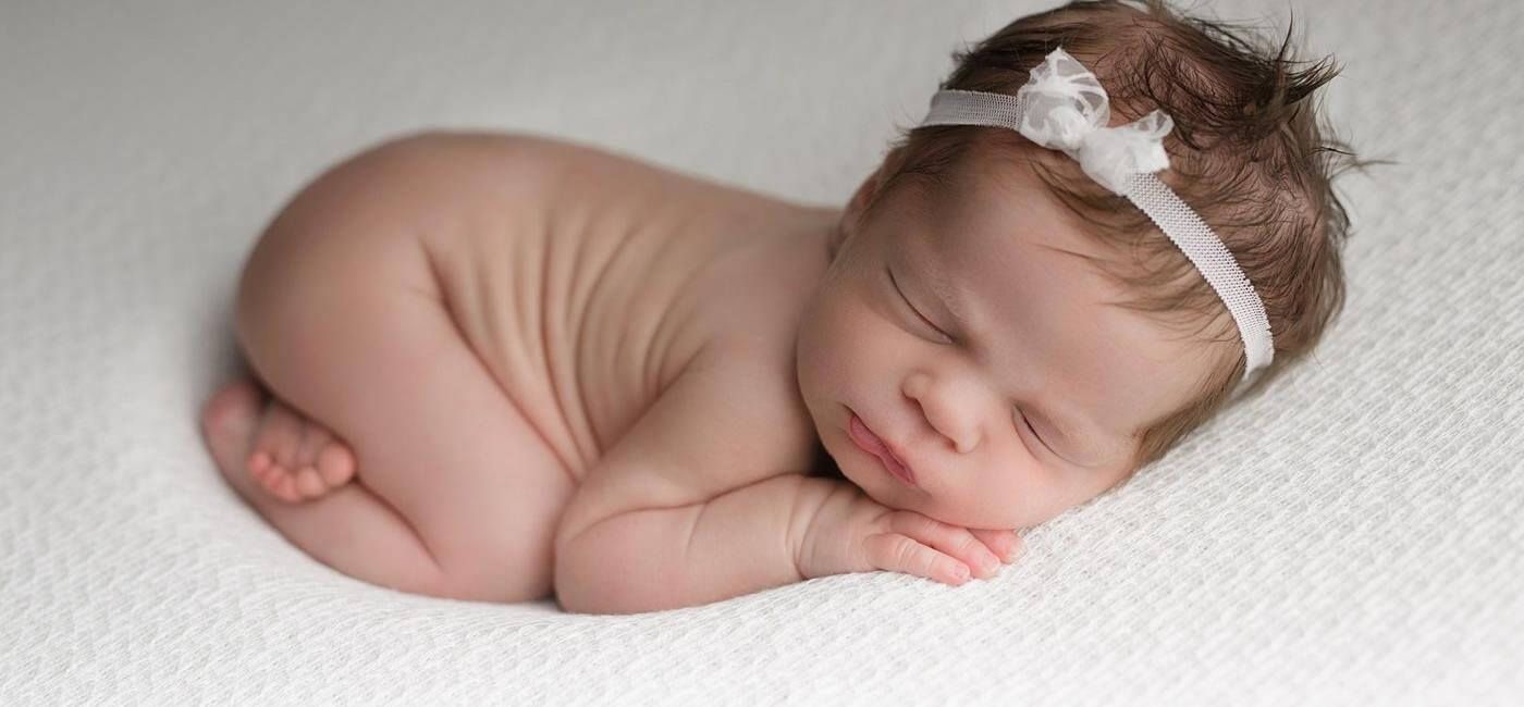 Sleeping newborn curled on a white blanket, wearing a white headband.