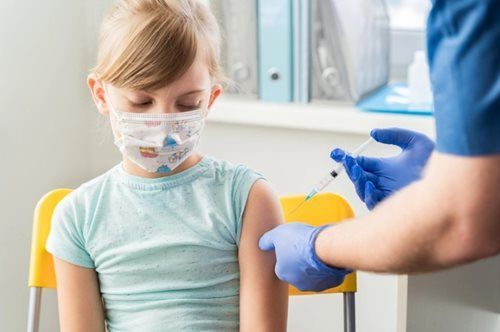 A little girl wearing a mask is getting an injection in her arm.