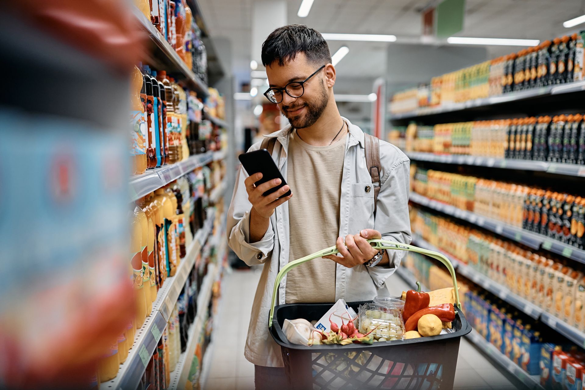 Man in glasses looks at phone, shopping in grocery store aisle, holding basket.