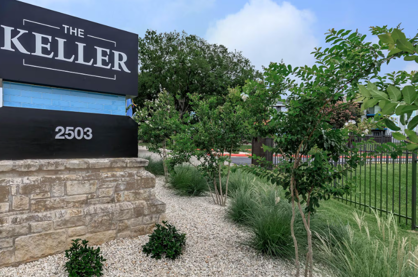 Stone sign for “The Keller” at 2503, with landscaped greenery and a black metal fence under a blue sky